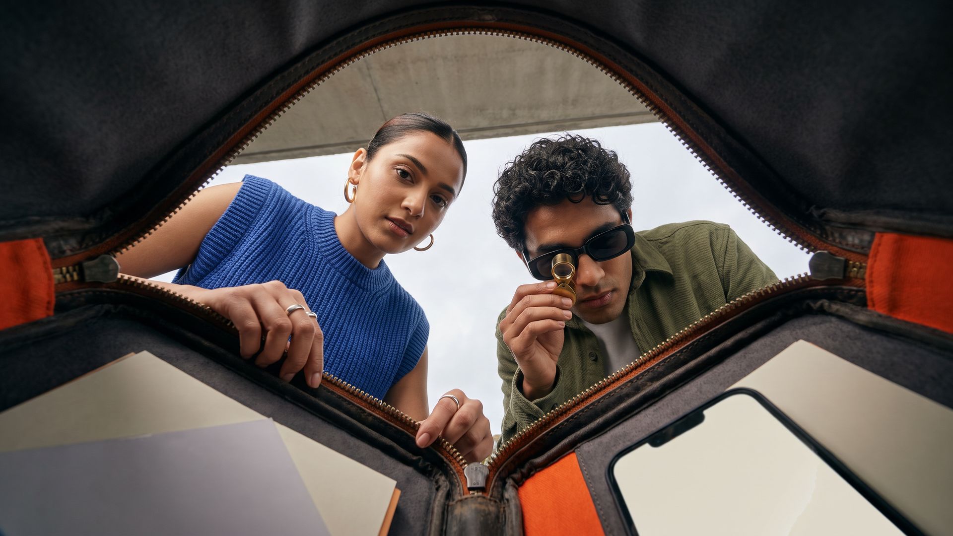 Two young Indian marketers unzip an orange-lined portfolio case and peer in, one holding a jeweller's loupe, seen from inside the case.