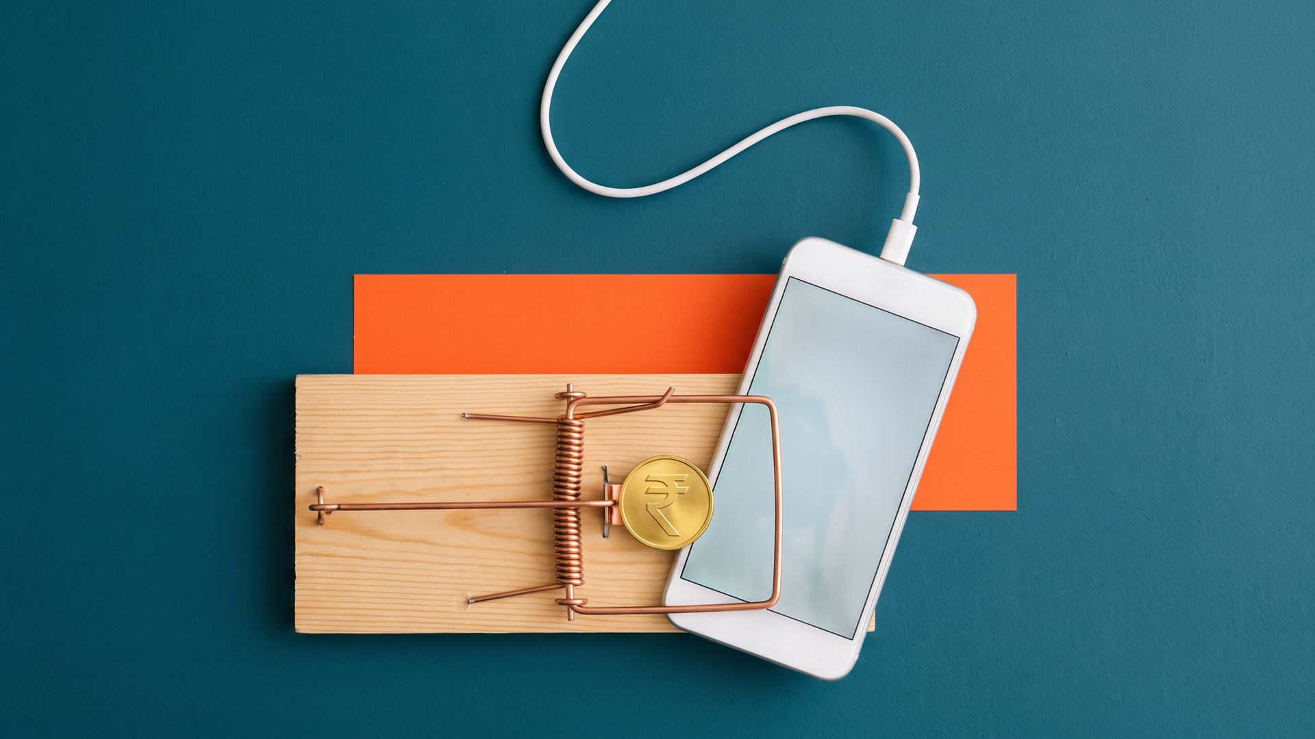 Overhead photo of a white smartphone caught in a wooden mousetrap baited with a rupee coin, its charging cable curling up like a tail on a teal and orange wall.